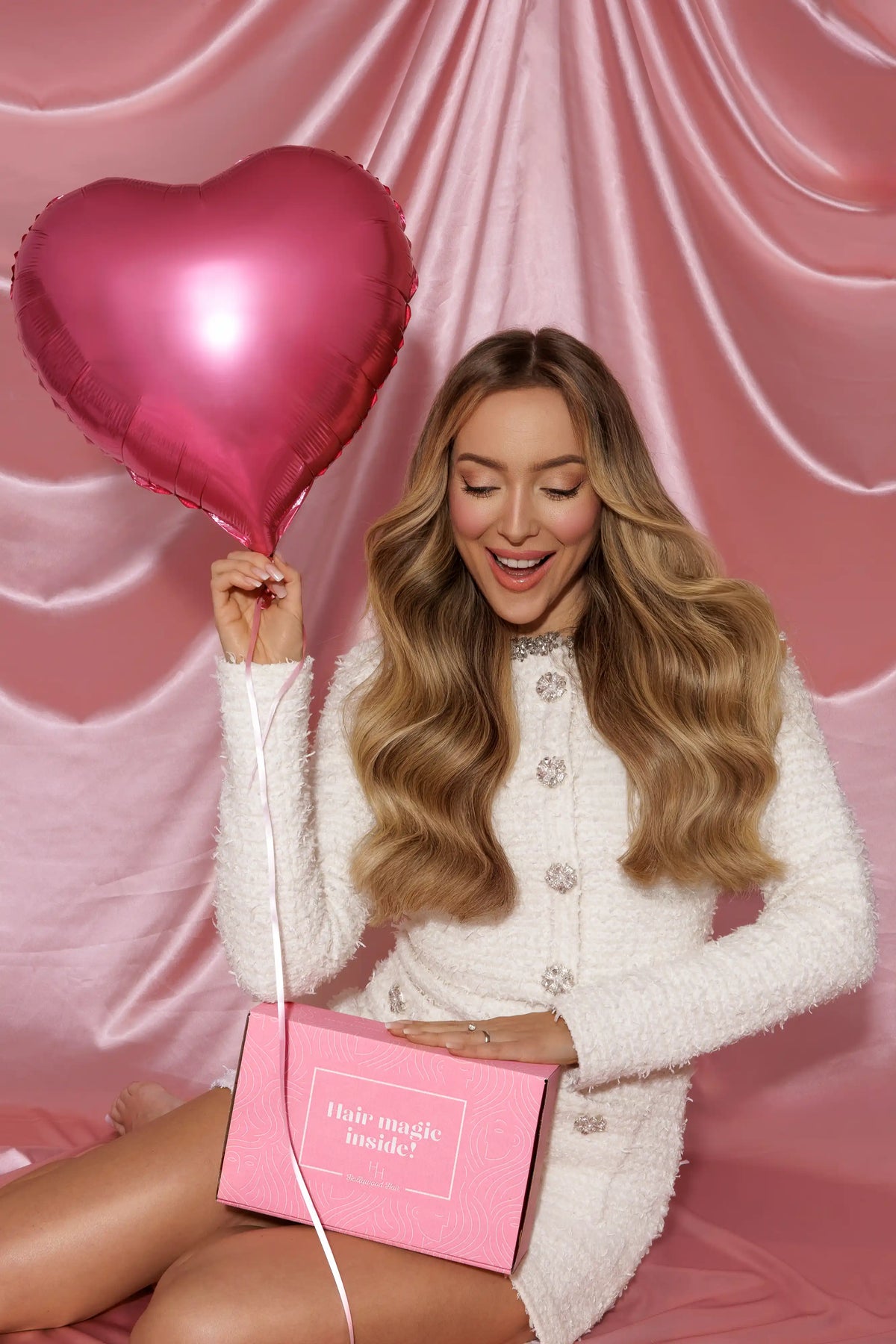 Uśmiechnięta kobieta trzyma różowy balon w kształcie serca oraz różowe pudełko z napisem, na tle jedwabistego różowego tła, co przywodzi na myśl radość i pozytywne emocje